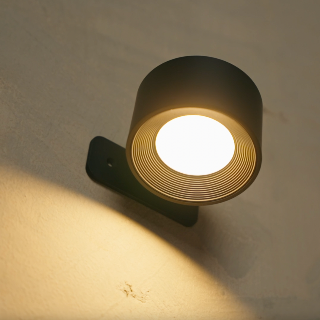 A sleek black wall-mounted lamp with a round, ribbed design, casting a warm golden light against a textured wall.
