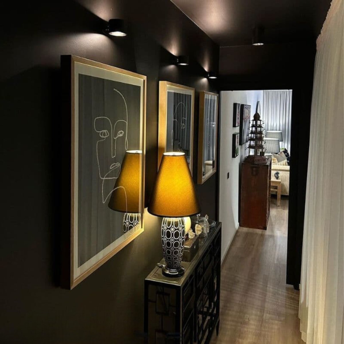 A stylish hallway featuring two abstract face artworks, decorative lamps, and a glimpse of a cozy living room in the background.