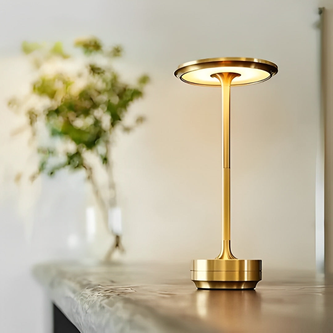 A stylish gold table lamp with a circular shade, casting warm light, placed on a smooth surface, with greenery softly blurred in the background.