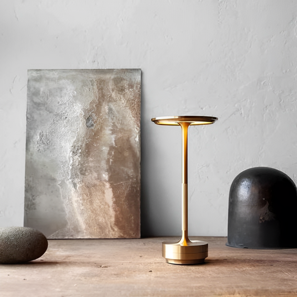 A stylish gold wireless table lamp stands on a wooden table beside a smooth gray stone and an abstract metallic art piece against a textured wall.