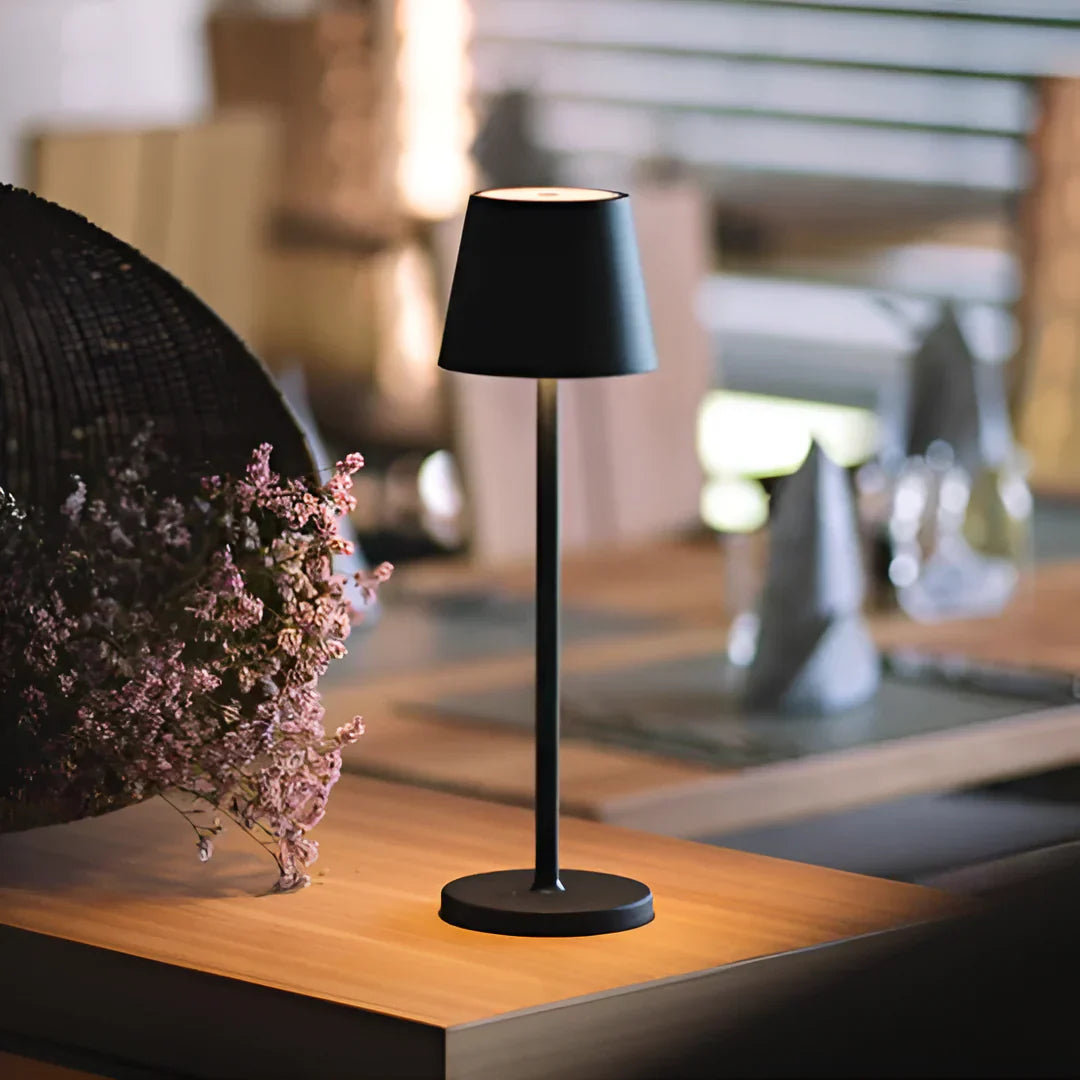 A sleek wireless black table lamp with a modern design, placed on a wooden table adorned with delicate pink flowers in a stylish setting.