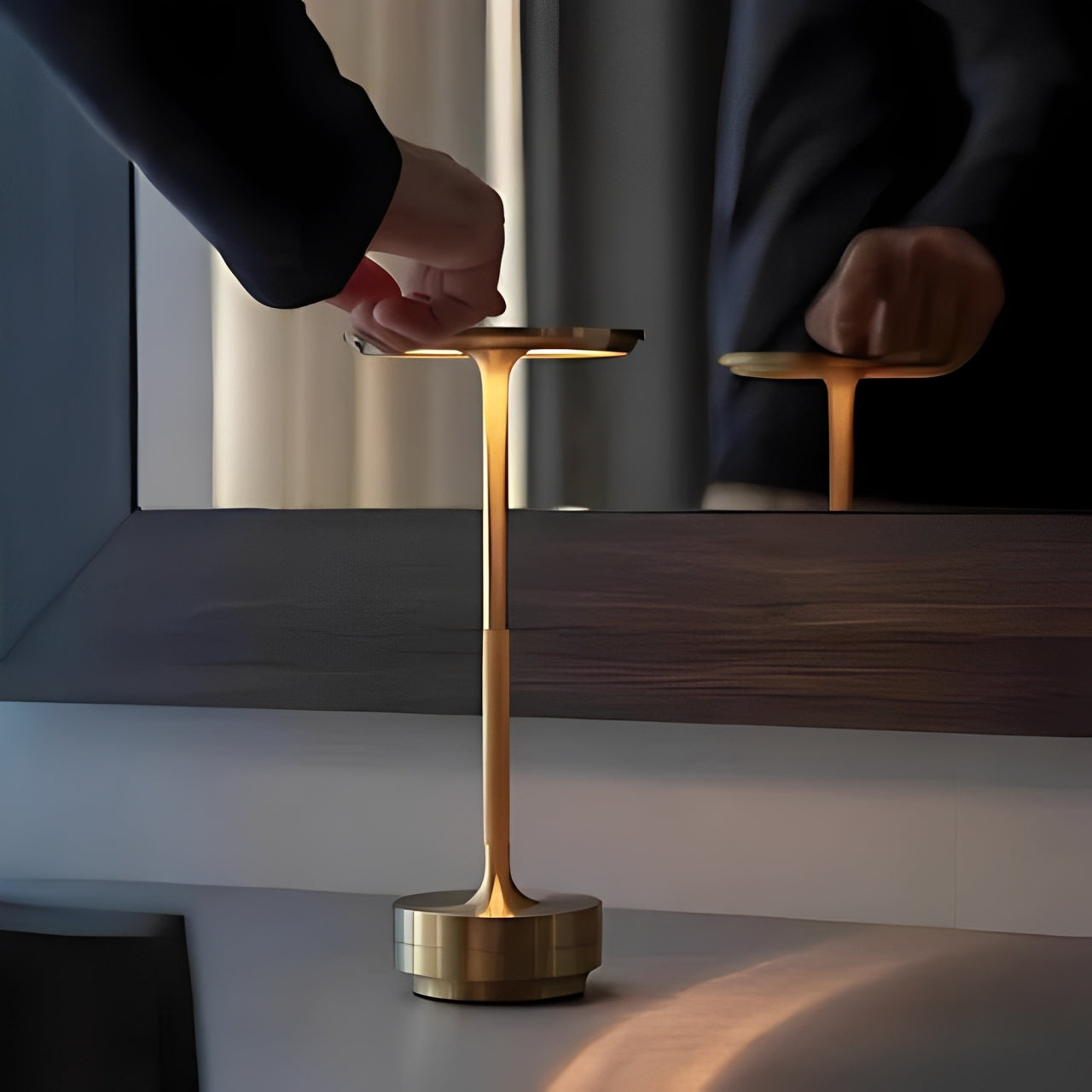 A hand adjusts a sleek, modern gold lamp on a desk, reflecting softly in the mirror behind it.