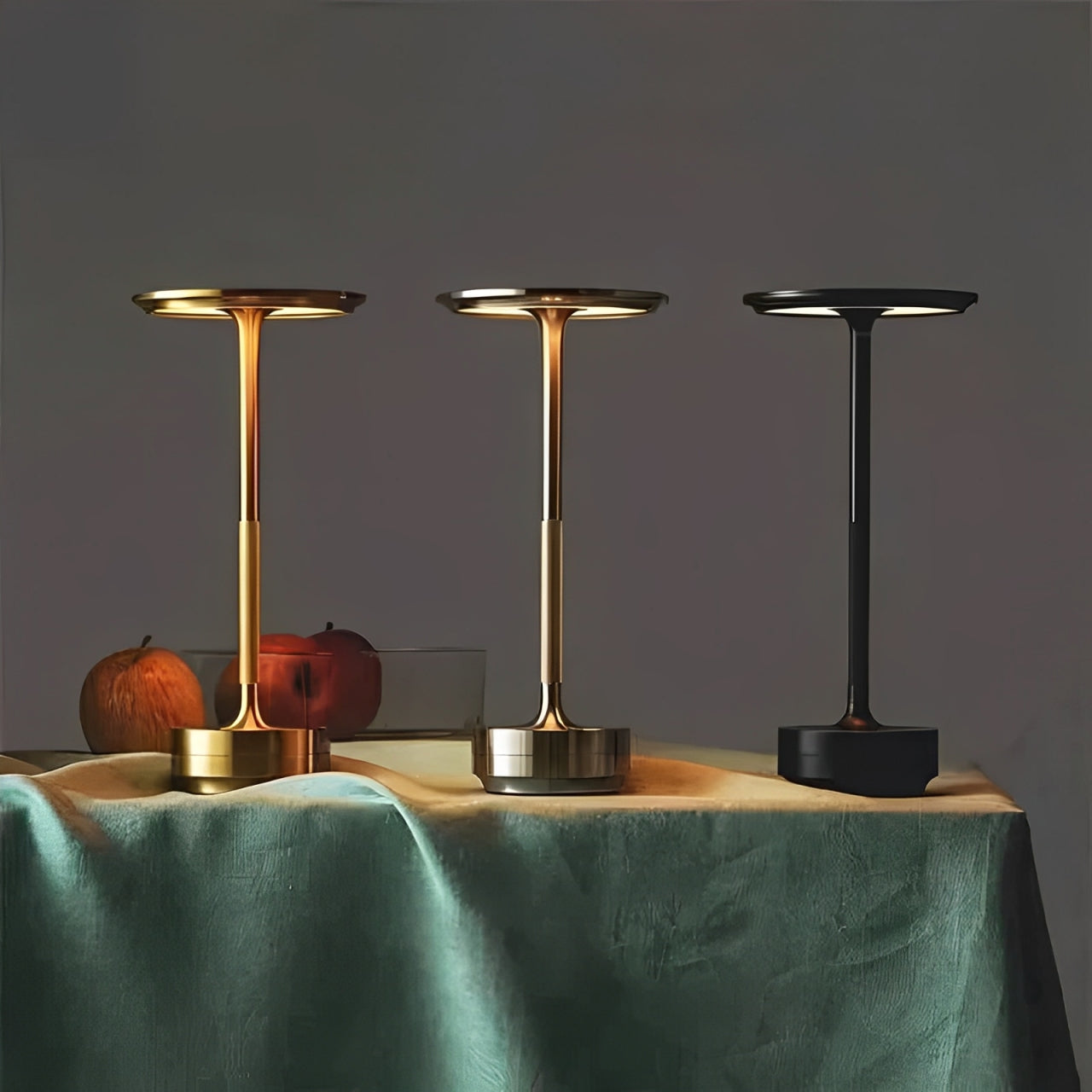 Three modern table lamps in gold, silver, and black, casting soft light on a green tablecloth beside decorative apples.