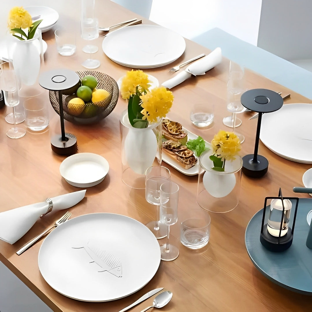 A beautifully set dining table with white plates, two modern table lamps, elegant glassware, yellow flowers, and a fruit bowl, creating a welcoming atmosphere.
