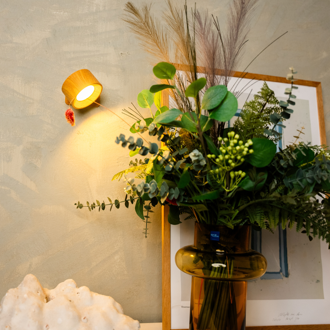 A warm wall lamp illuminates a lush green bouquet in a glass vase, framed art, and a textured wall, creating a cozy ambiance.