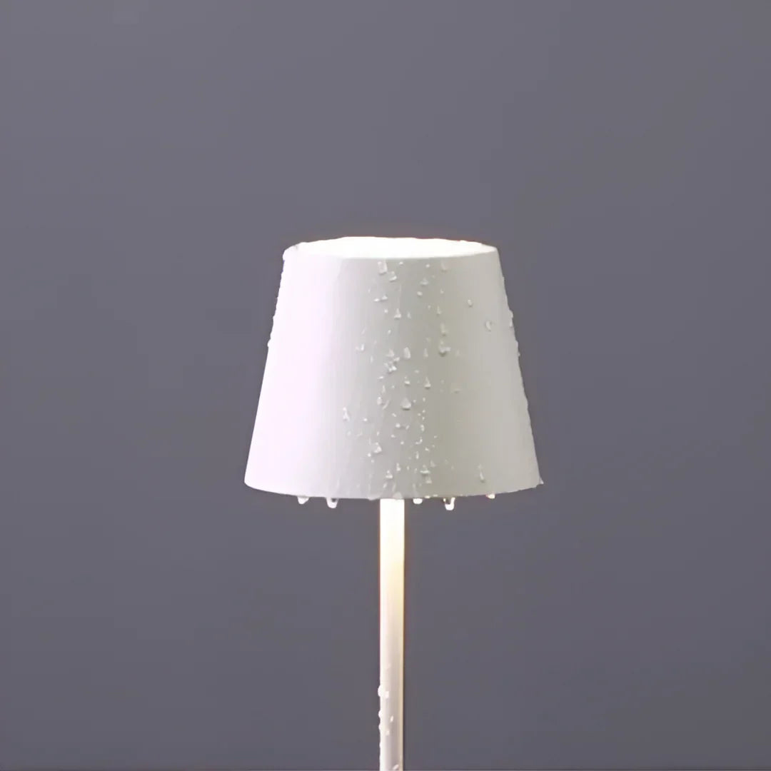 A sleek, waterproof white cordless lamp with a wet shade, featuring droplets, stands against a gray backdrop, radiating soft light from its base.