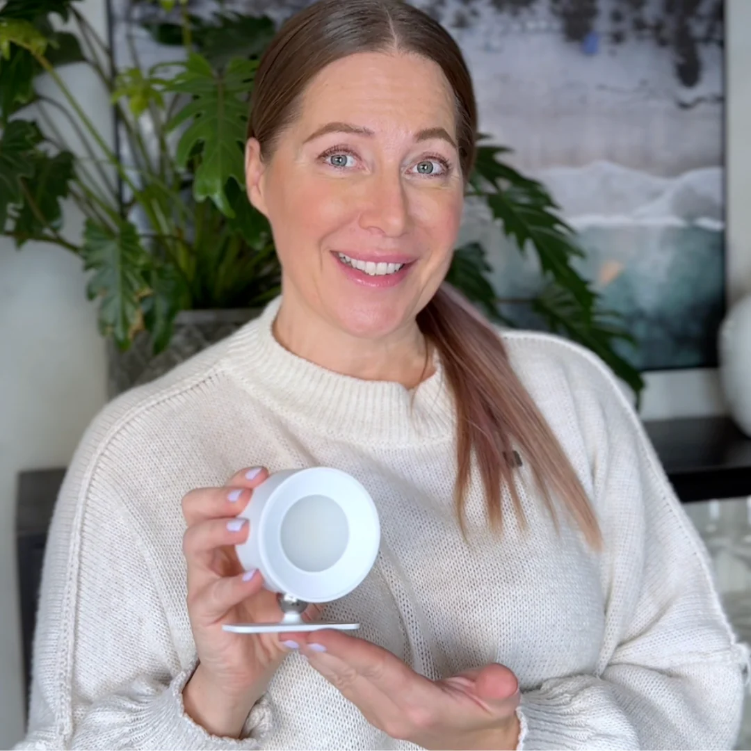 Woman holding a white wall lamp in an indoor setting with plants and a painting in the background
