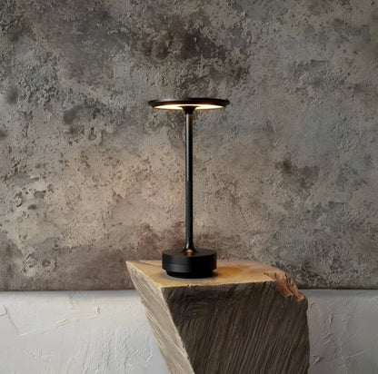 A sleek black table lamp stands on a wooden block against a textured gray wall, exuding a modern and minimalist aesthetic.