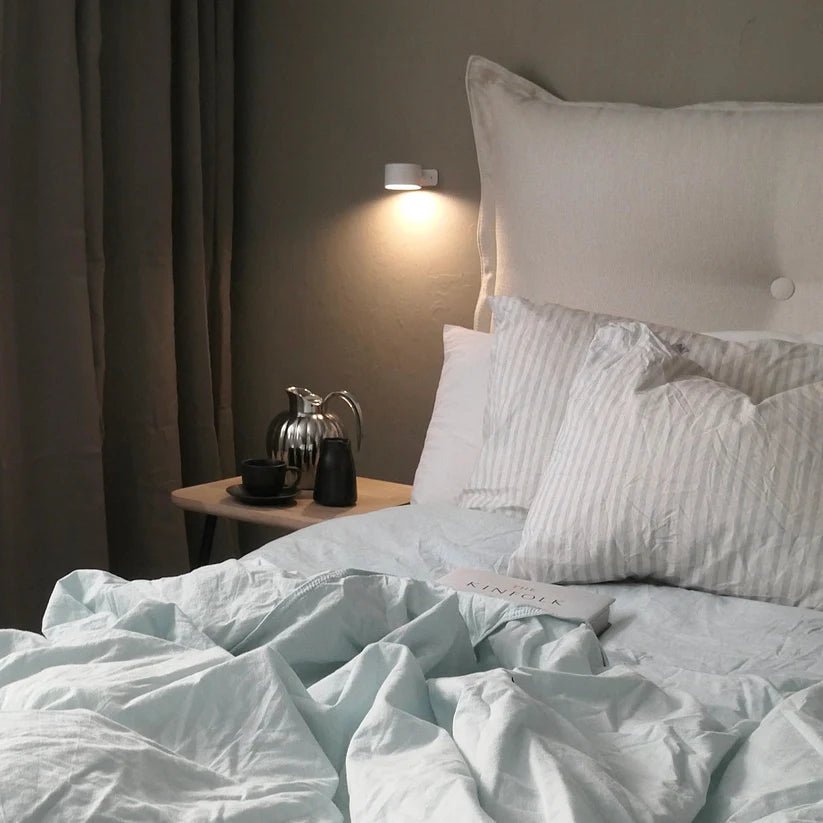 Cozy bed with rumpled blue linens, plush pillows, and a bedside table featuring a coffee set and a book under soft, wireless wall lamp lighting.