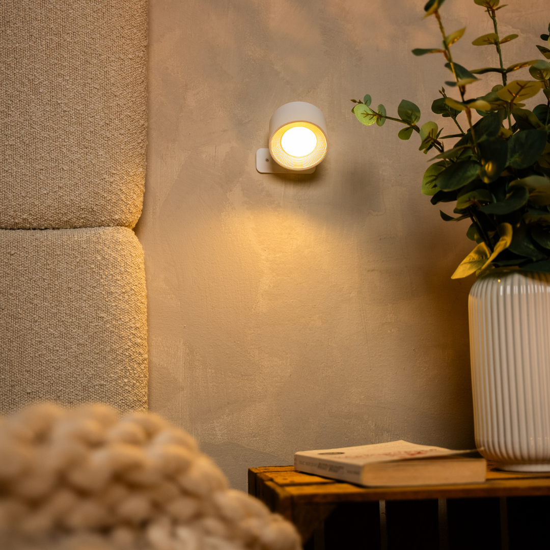 A cozy corner featuring a wall-mounted lamp, a textured headboard, a vase with greenery, and a stack of books on a wooden table.