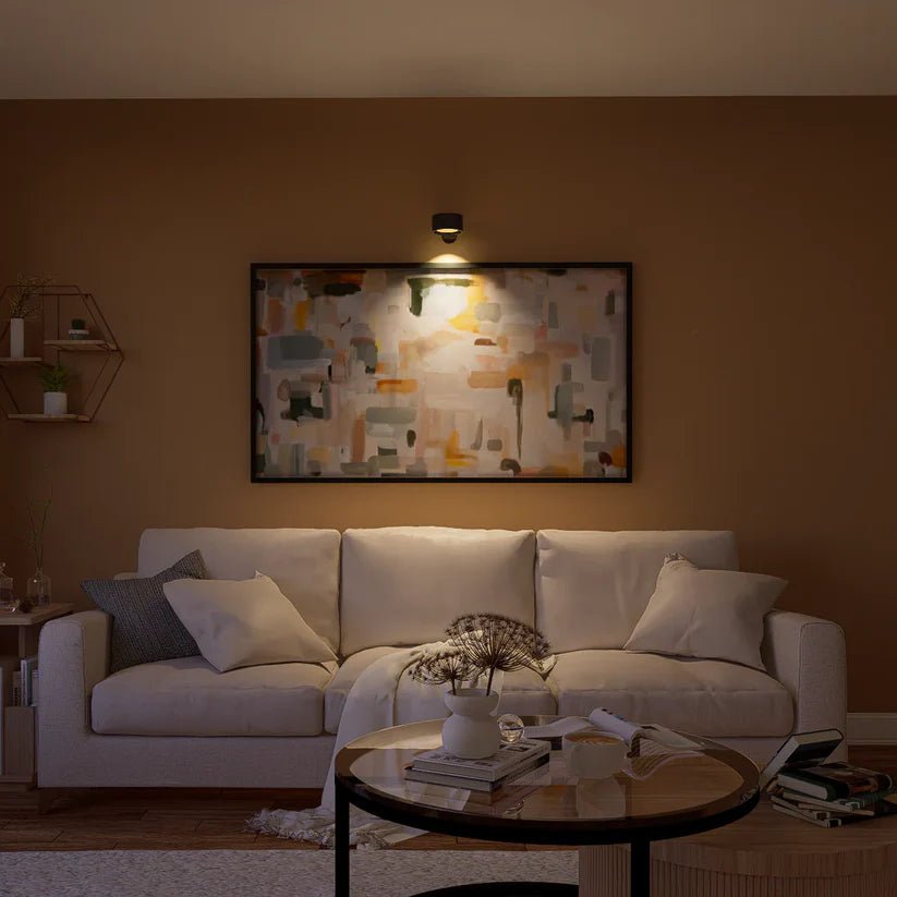 Cozy living room with a white sofa, round table, and abstract wall art illuminated by a wall light, against a warm brown backdrop.