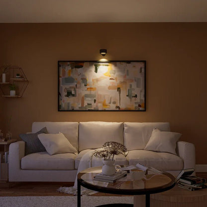Cozy living room with a white sofa, round table, and abstract wall art illuminated by a wall light, against a warm brown backdrop.