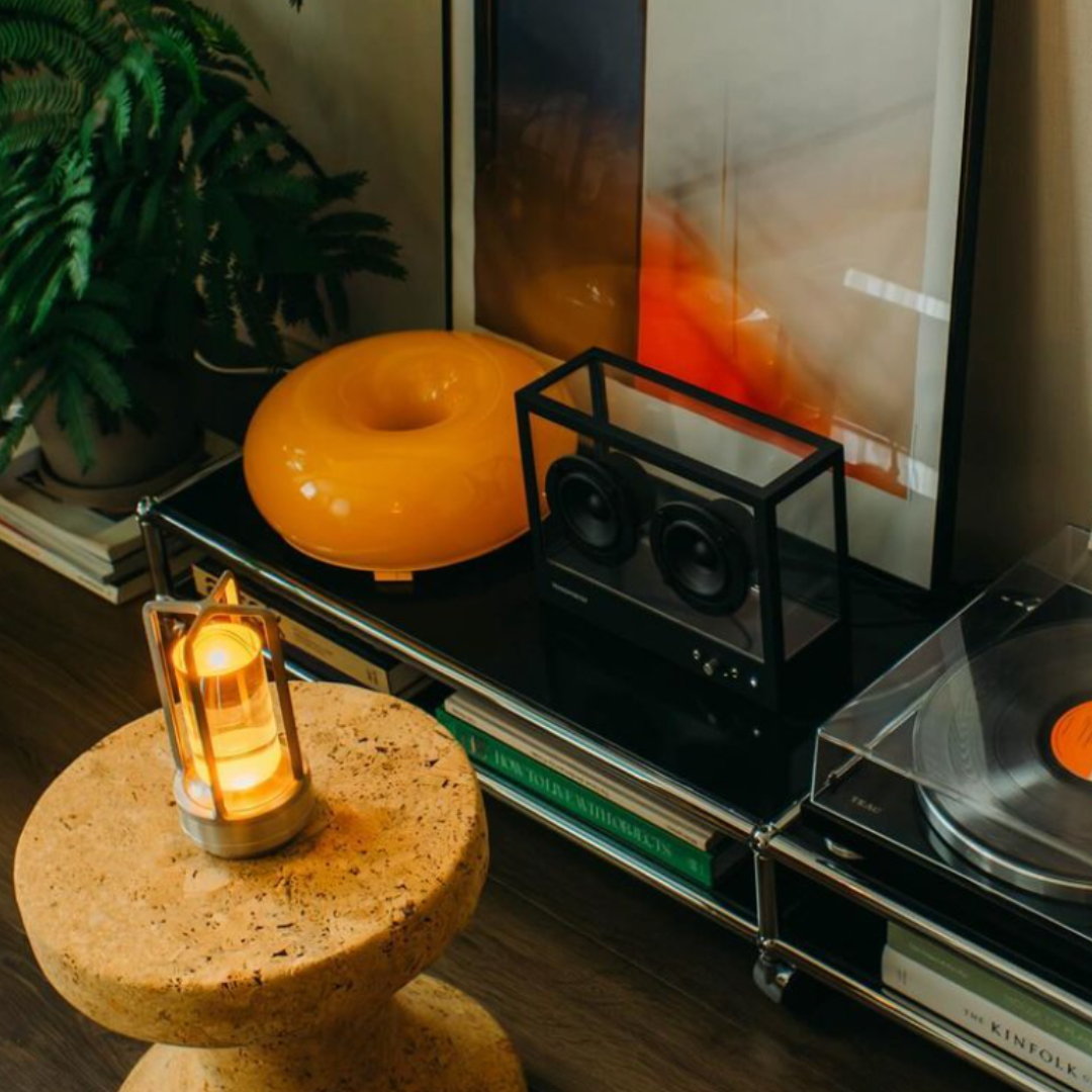 A cozy living space featuring a cork stool, table lamp, black speakers, and a record player atop a stylish shelf.