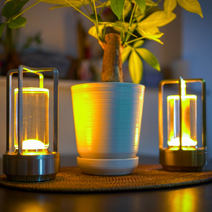 Two cordless glowing table lantern lamps flank a bright white pot containing a leafy green plant, all set atop a textured brown placemat.