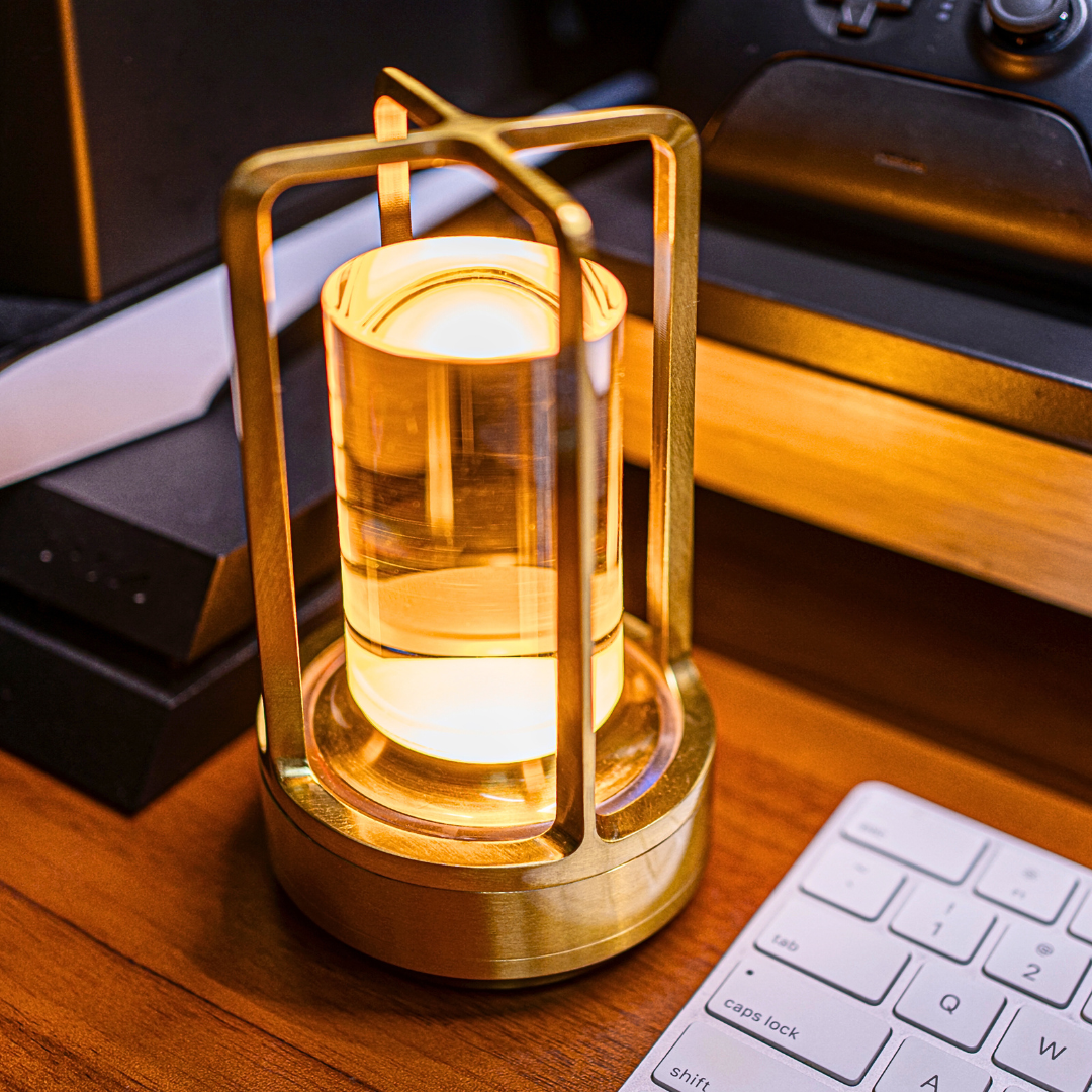 A gold and glass table lamp emits a warm light on a wooden desk next to a white computer keyboard and electronic devices.