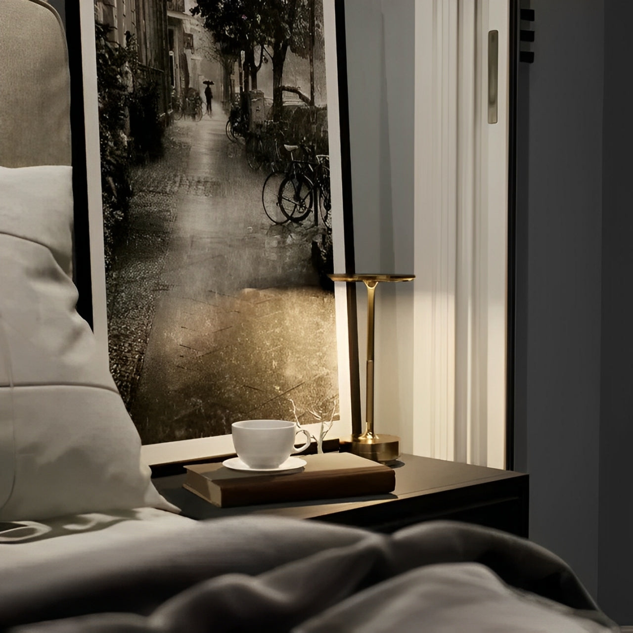 Cozy bedside scene featuring a gold lamp, a cup on a saucer, a book, and a vintage black and white photograph of a rain-soaked street.