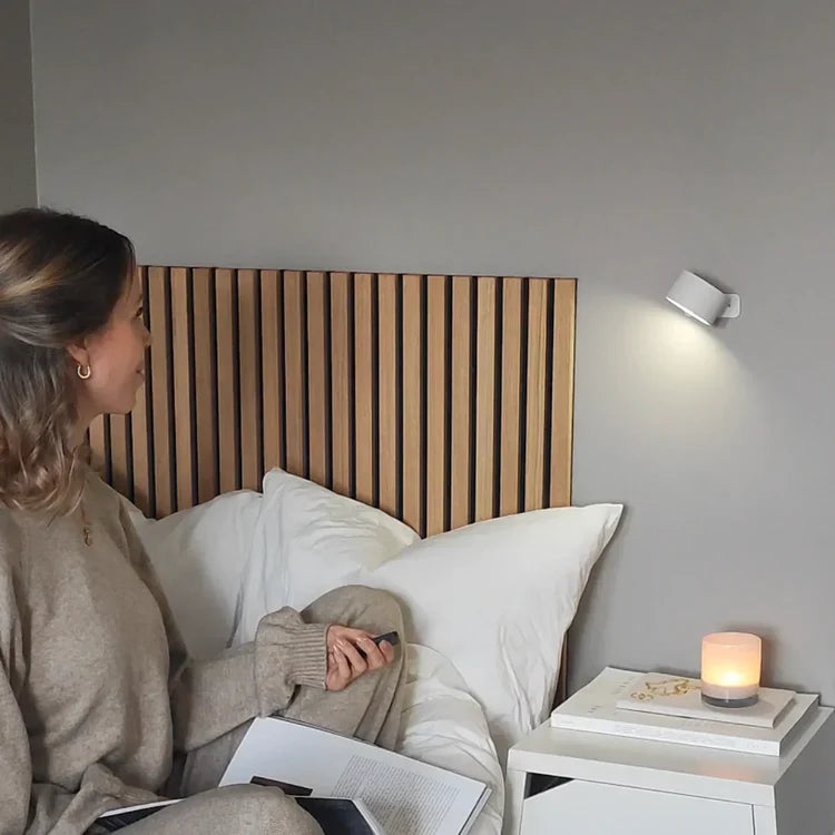 Woman sitting on a bed with a remote control, reading a book, next to a nightstand with a lit candle and white wall lamp.