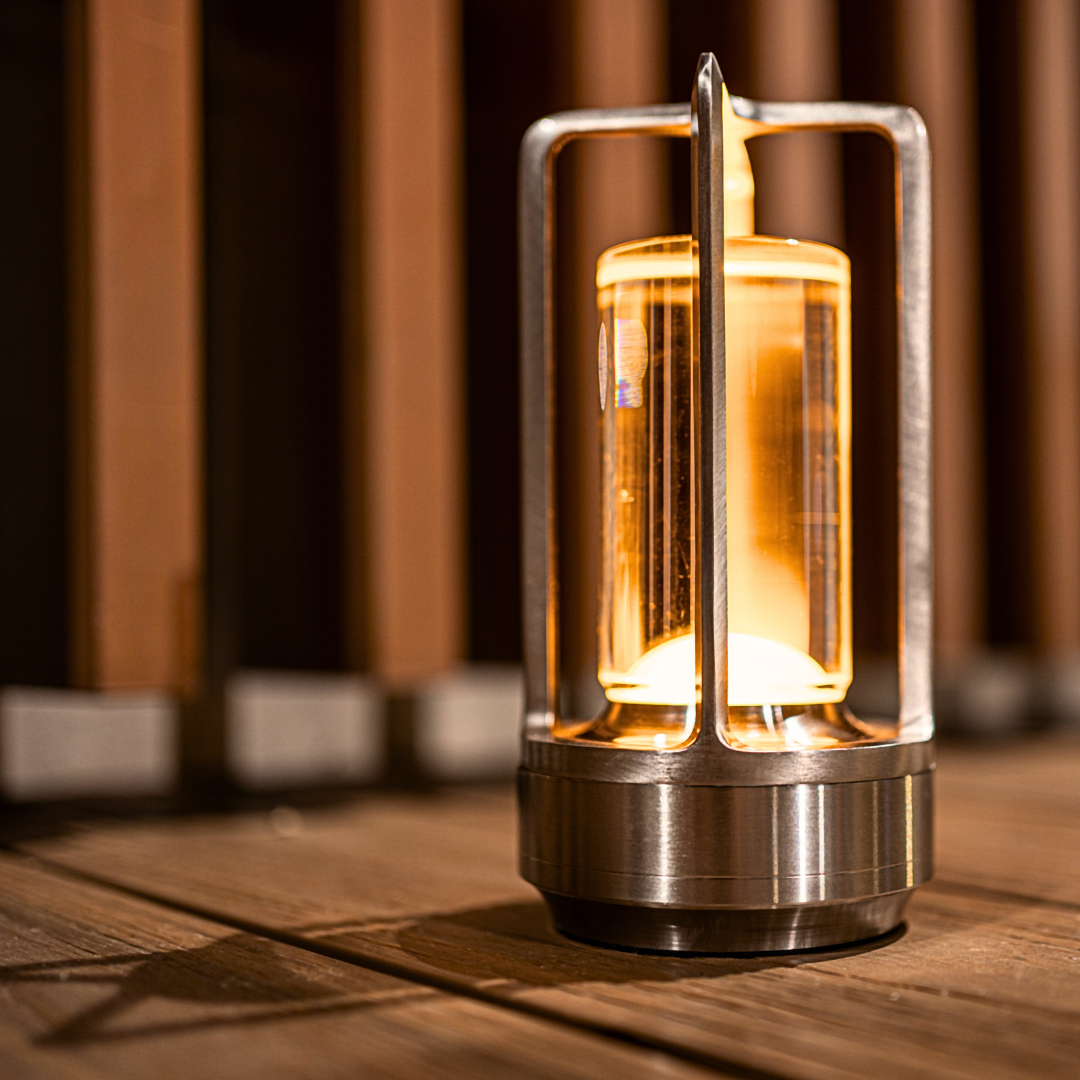 A stylish metal lantern table lamp with a warm glowing bulb, resting on wooden decking, against a blurred wooden backdrop.