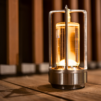 A stylish metal lantern table lamp with a warm glowing bulb, resting on wooden decking, against a blurred wooden backdrop.