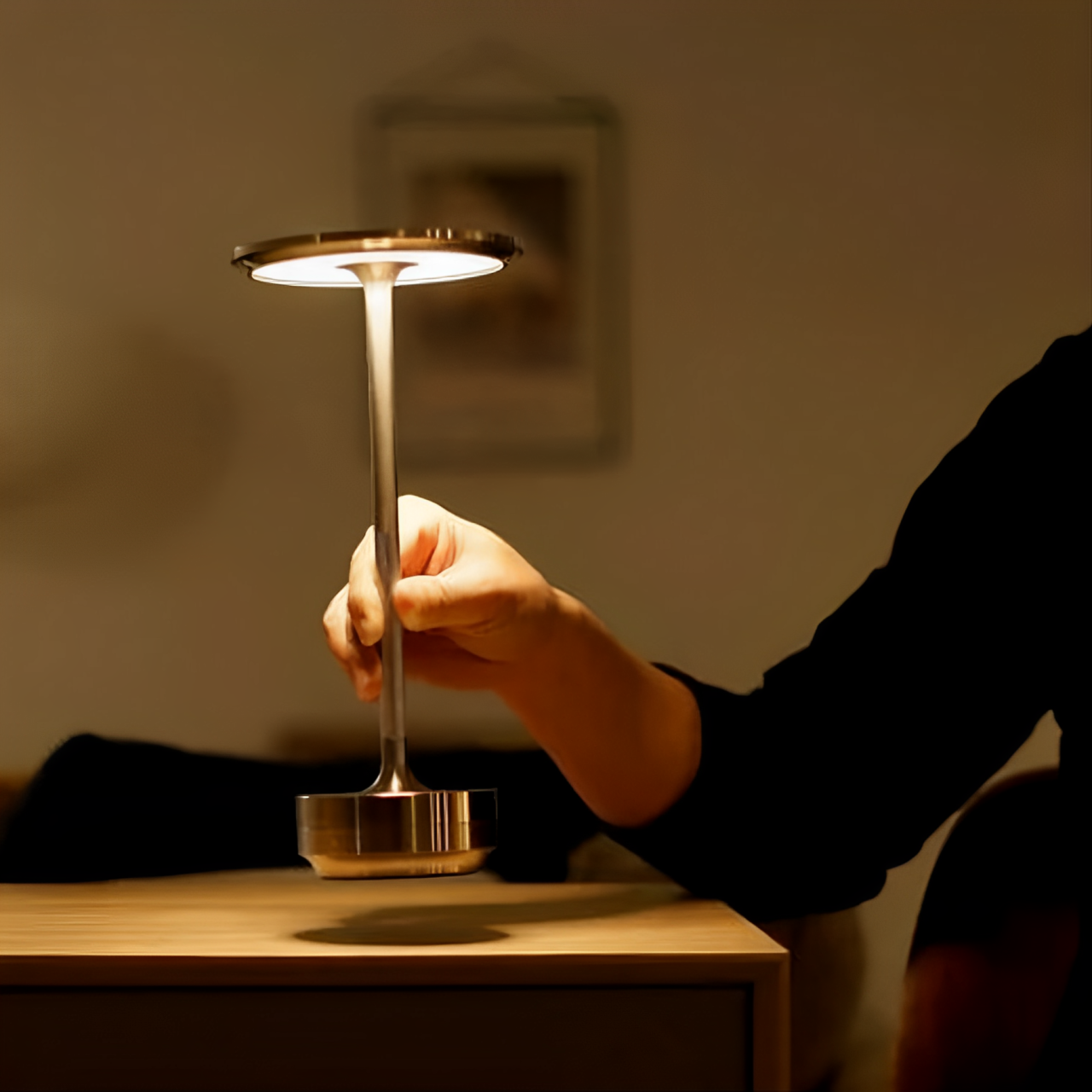 A hand reaches for a sleek, modern lamp with a circular head, illuminating a cozy, dimly lit room with warm light.