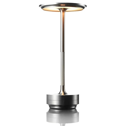 A sleek, modern silver table lamp with a circular top and slender pole, reflecting softly on a surface below.