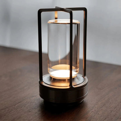 A modern, cylindrical table lamp with a glass center and metal frame sits on a wooden surface, emitting a warm light.