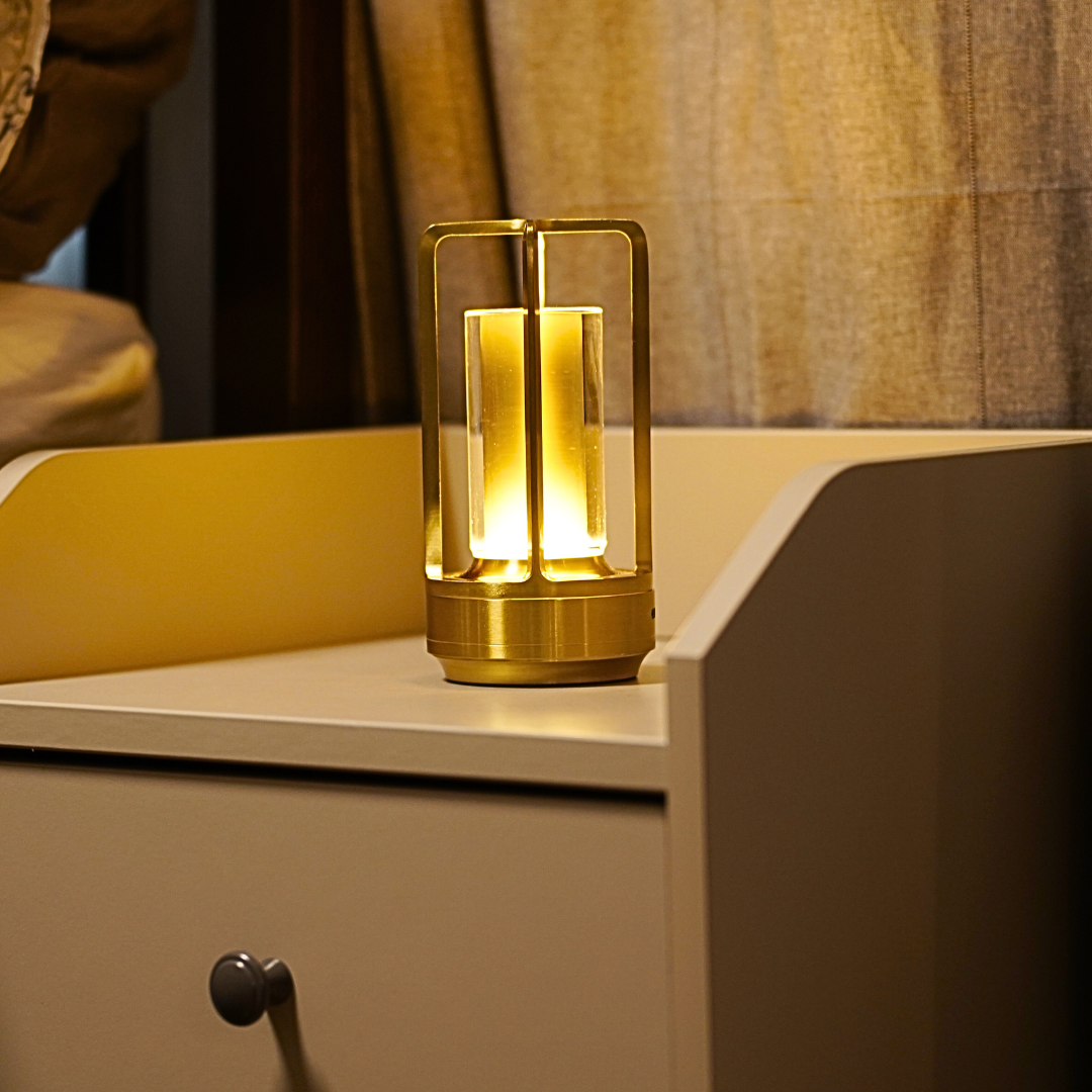 A stylish gold lantern-style lamp emits a warm glow, placed on a beige nightstand beside softly draped curtains.