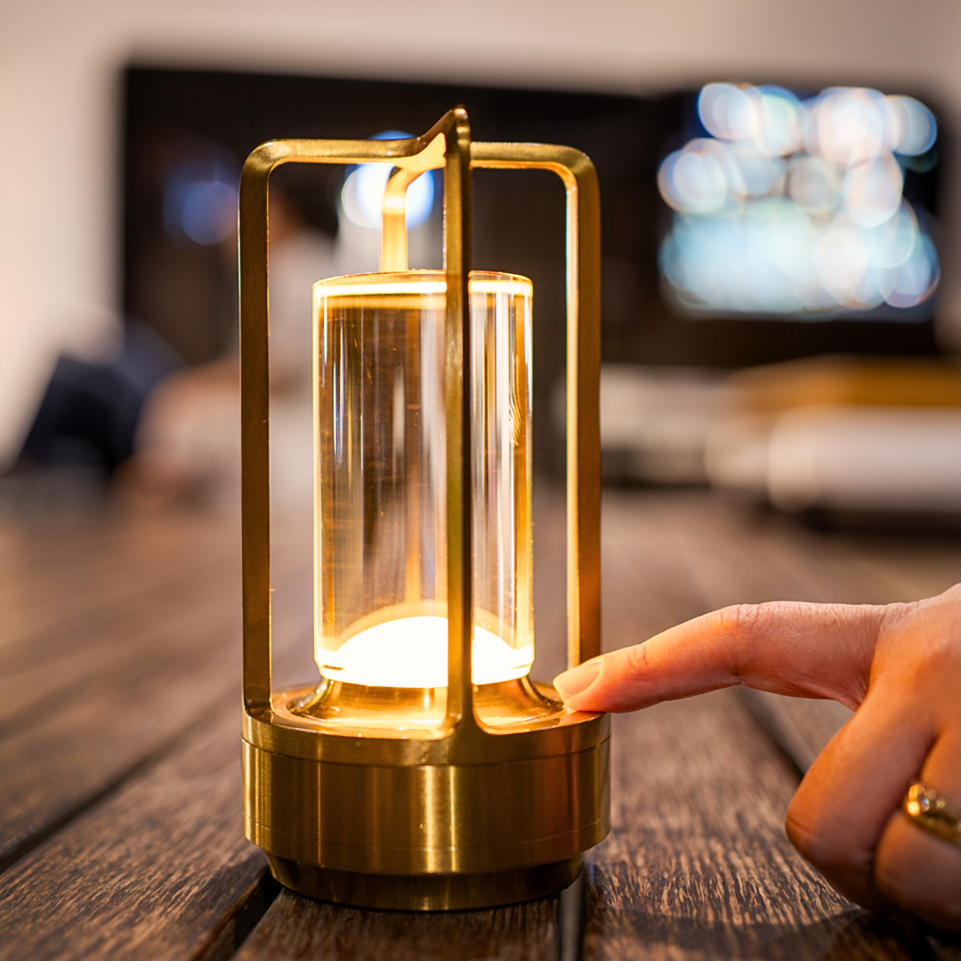 A person’s finger touches a stylish table lantern, illuminating its clear cylindrical glass core on a wooden table.