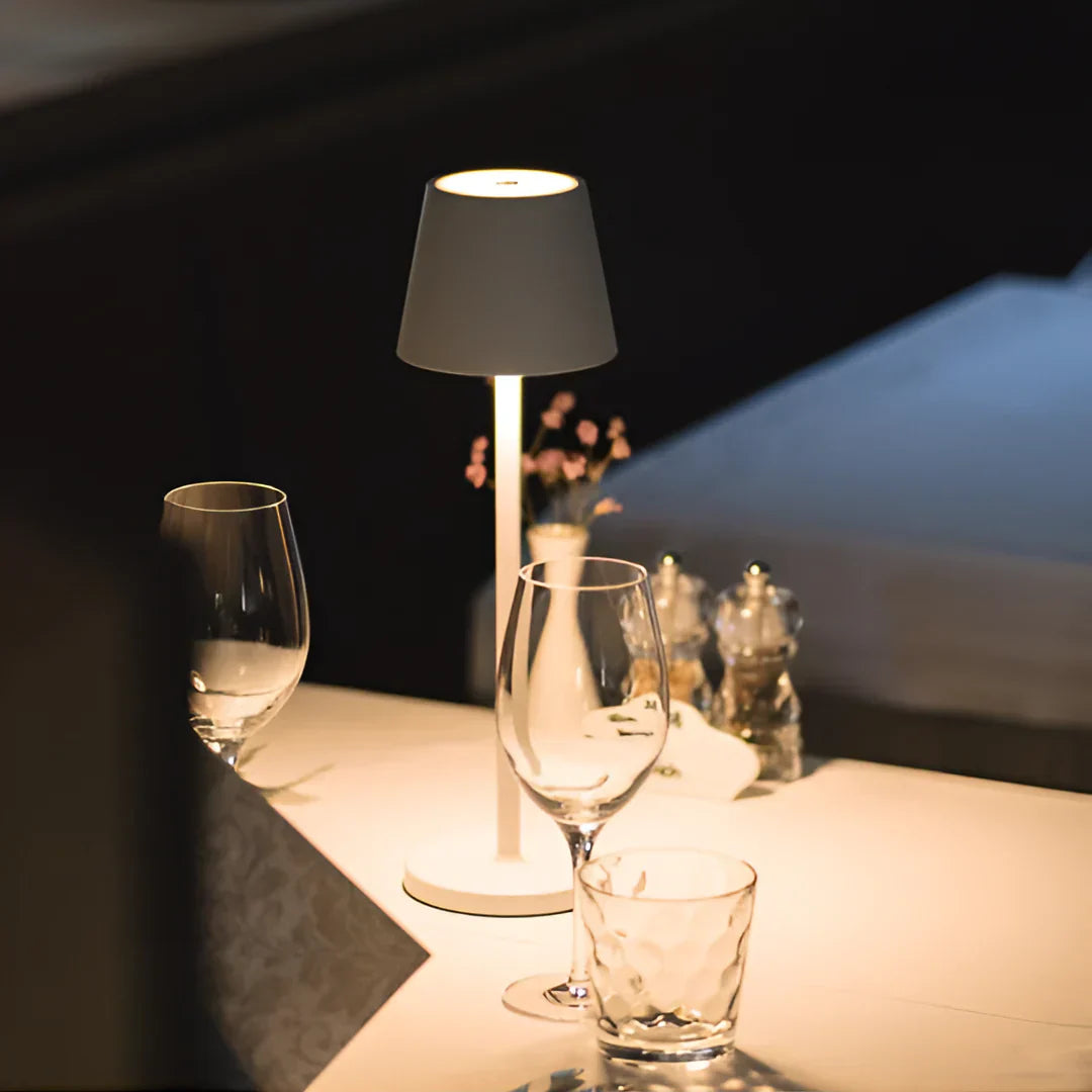 A table lamp illuminates a restaurant table set with empty glasses, salt and pepper shakers, and a white tablecloth.