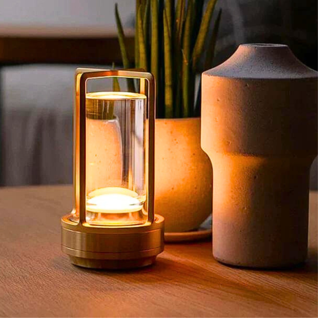 An elegant glass lantern table lamp with a warm glow, placed on a wooden table next to textured ceramic vases and greenery.