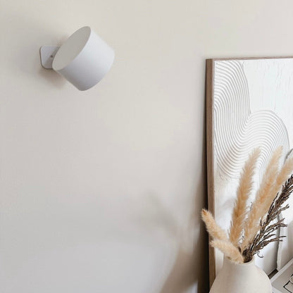 A modern white wall lamp illuminates a beige wall beside a textured mirror and a vase with dried pampas grass.