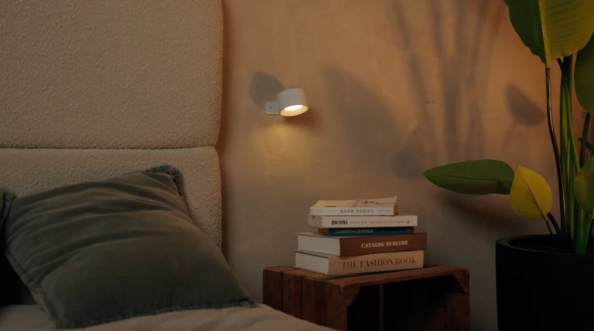 Stack of books on a wooden nightstand next to a bed with a green pillow, illuminated by a wall light.