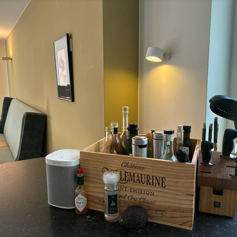 A wooden crate filled with assorted bottles and kitchen utensils sits on a dark countertop, illuminated by a nearby wall light.