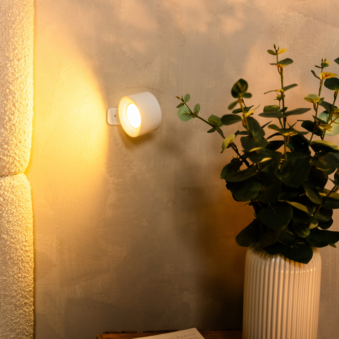 A wireless white wall lamp casts a warm glow on a textured wall beside a green plant in a ribbed white vase, creating a cozy atmosphere.