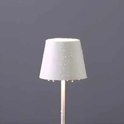 A sleek, waterproof white cordless lamp with a wet shade, featuring droplets, stands against a gray backdrop, radiating soft light from its base.